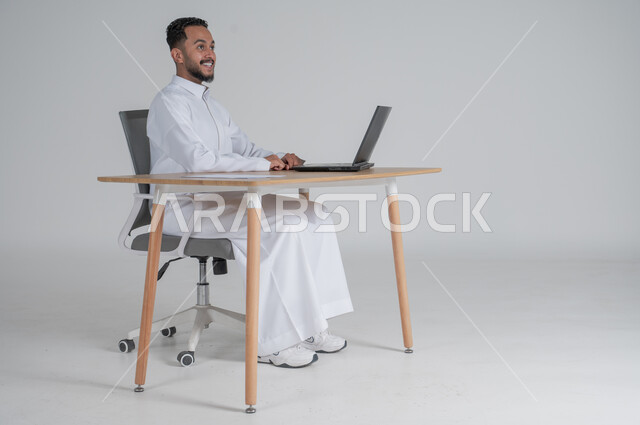 استخدام التقنيات الحديثة والمتطورة، بورتريه لشاب عربي سعودي خليجي يرتدي الثوب التقليدي و يعمل عن بعد لاستخدام الحاسوب المحمول ينظر الى شيء ما بإيماءات السعادة و الفرح، إجراء مكالمة فيديو عبر جهاز الكمبيوتر، بيئة العمل السعودي، خلفية بيضاء