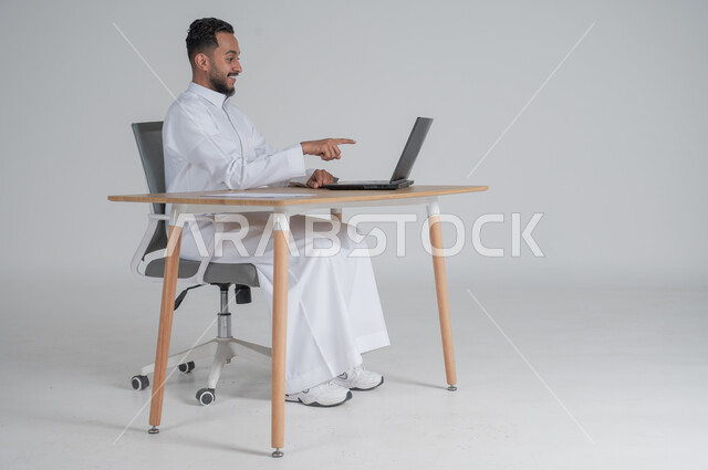 استخدام التقنيات الحديثة والمتطورة، بورتريه لشاب عربي سعودي خليجي يرتدي الثوب التقليدي و يعمل عن بعد يشير بأصبعه السبابة الى الحاسوب المحمول بإيماءات السعادة و الفرح، إجراء مكالمة فيديو عبر جهاز الكمبيوتر، بيئة العمل السعودي، خلفية بيضاء