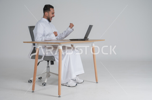 استخدام التقنيات الحديثة والمتطورة، بورتريه لشاب عربي سعودي خليجي يرتدي الثوب التقليدي و يعمل عن بعد باستخدام الحاسوب المحمول يرفع قبضة يديه بإيماءات الفوز والسعادة والانتصار، إجراء مكالمة فيديو عبر جهاز الكمبيوتر، بيئة العمل السعودي، خلفية بيضاء