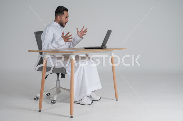استخدام التقنيات الحديثة والمتطورة، بورتريه لشاب عربي سعودي خليجي يرتدي الثوب التقليدي و يعمل عن بعد باستخدام الحاسوب المحمول بإيماءات الغضب و الصراخ، إجراء مكالمة فيديو عبر جهاز الكمبيوتر، بيئة العمل السعودي، خلفية بيضاء