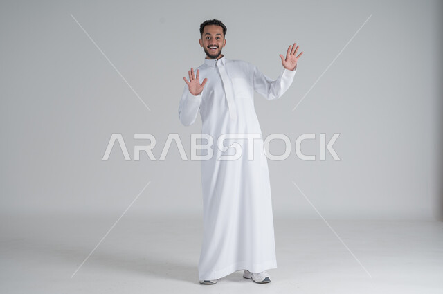 الشعور بالدهشة والتعجب والاستغراب، بورتريه لشاب عربي خليجي سعودي يرتدي الزي التقليدي و يرفع يديه للأعلى بتعابير المفاجأة, النظر إلى الكاميرا بإيماءات الفرح والسرور , صورة الجسم بالكامل, خلفية بيضاء