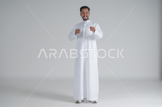 النظر الى الكاميرا بسخرية والاستهزاء، بورتريه لرجل عربي خليجي سعودي يرتدي الثوب التقليدي يرفع يديه للأعلى بإيماءات التعجب والفرح، الجسم بالطول الكامل، خلفية بيضاء