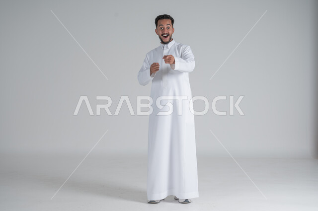 النظر الى الكاميرا بسخرية والاستهزاء، بورتريه لرجل عربي خليجي سعودي يرتدي الثوب التقليدي يرفع يديه للأعلى بإيماءات التعجب والفرح، الجسم بالطول الكامل، خلفية بيضاء
