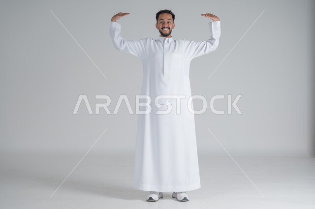 النظر الى الكاميرا بسعادة و سرور، بورتريه لرجل عربي خليجي سعودي مبتسم يرتدي الثوب التقليدي يرفع يديه للأعلى بايماءات اظهار حجم شيء ما، الجسم بالطول الكامل، خلفية بيضاء