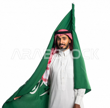 بورتريه لرجل عربي خليجي سعودي يضع على ظهره علم المملكة العربية السعودية، يبتسم إبتسامة هوليود، ذكرى اليوم الوطني السعودي، الاحتفال بالعيد الوطني السعودي، خلفية بيضاء لرجل سعودي يرتدي زي تقليدي سعودي مع شماغ وعقال