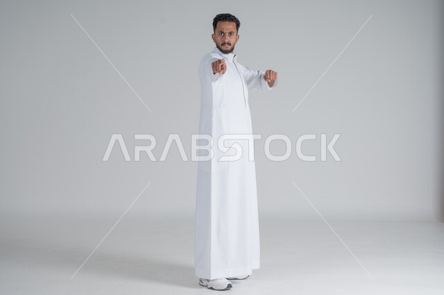 تعابير الجدية و الحزم, الاتهام و إلقاء اللوم, الإشارة إلى شيء ما, بورتريه لرجل عربي سعودي خليجي يرتدي الثوب التقليدي ينظر إلى الكاميرا بإيماءات الغضب, الجسم بالطول الكامل, خلفية بيضاء