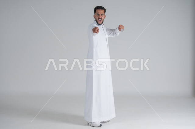 تعابير الجدية و الحزم, الاتهام و إلقاء اللوم, الإشارة إلى شيء ما, بورتريه لرجل عربي سعودي خليجي يرتدي الثوب التقليدي ينظر إلى الكاميرا بإيماءات الغضب, الجسم بالطول الكامل, خلفية بيضاء