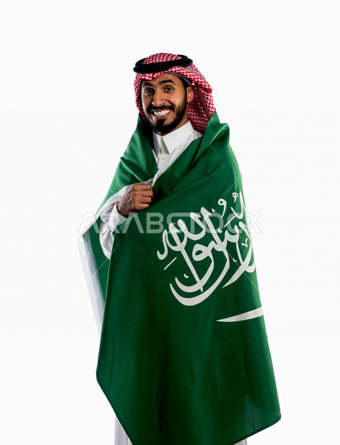 بورتريه لرجل عربي خليجي سعودي يضع على ظهره علم المملكة العربية السعودية، يبتسم إبتسامة هوليود، ذكرى اليوم الوطني السعودي، الاحتفال بالعيد الوطني السعودي، خلفية بيضاء لرجل سعودي يرتدي زي تقليدي سعودي مع شماغ وعقال