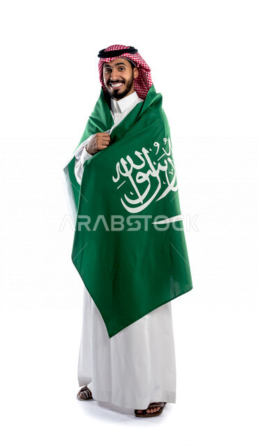 بورتريه لرجل عربي خليجي سعودي يضع على ظهره علم المملكة العربية السعودية، يبتسم إبتسامة هوليود، ذكرى اليوم الوطني السعودي، الاحتفال بالعيد الوطني السعودي، خلفية بيضاء لرجل سعودي يرتدي زي تقليدي سعودي مع شماغ وعقال
