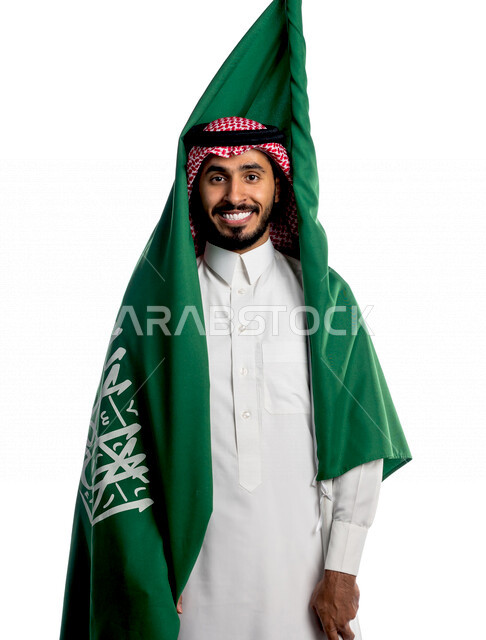 بورتريه لرجل عربي خليجي سعودي يضع على ظهره علم المملكة العربية السعودية، يبتسم إبتسامة هوليود، ذكرى اليوم الوطني السعودي، الاحتفال بالعيد الوطني السعودي، خلفية بيضاء لرجل سعودي يرتدي زي تقليدي سعودي مع شماغ وعقال
