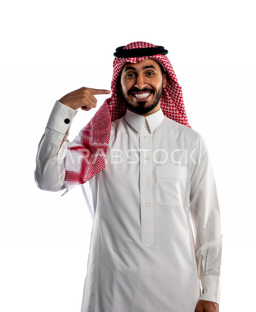 بورتريه لرجل سعودي خليجي مبتسم يقوم بجلسة تصوير، ينظر إلى الكاميرا، يشير بإصبعه على أسنانه بإيماءات وجه ويدين مختلفة ، إبتسامة هوليود ، أسنان جميلة ذات بياض ناصع، خلفية بيضاء لرجل سعودي مسلم يرتدي زي تقليدي سعودي مع شماغ وعقال