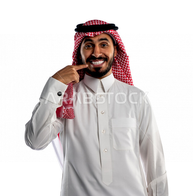 بورتريه لرجل سعودي خليجي مبتسم يقوم بجلسة تصوير، ينظر إلى الكاميرا، يشير بإصبعه على أسنانه بإيماءات وجه ويدين مختلفة ، إبتسامة هوليود ، أسنان جميلة ذات بياض ناصع، خلفية بيضاء لرجل سعودي مسلم يرتدي زي تقليدي سعودي مع شماغ وعقال