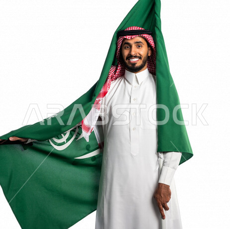 بورتريه لرجل عربي خليجي سعودي يضع على ظهره علم المملكة العربية السعودية، يبتسم إبتسامة هوليود، ذكرى اليوم الوطني السعودي، الاحتفال بالعيد الوطني السعودي، خلفية بيضاء لرجل سعودي يرتدي زي تقليدي سعودي مع شماغ وعقال