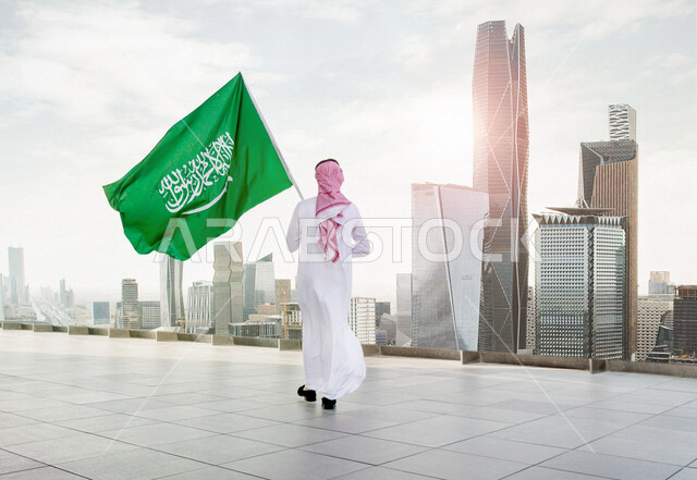 زاوية من الخلف لرجل عربي سعودي خليجي يرتدي الزي السعودي التقليدي مع شماغ وعقال، يحمل بيده علم السعودية مع خلفية لأبراج المملكة العربية السعودية وناطحات السحاب، الاحتفال باليوم الوطني السعودي، علم السعودية يرفرف في الأعلى بالسماء الزرقاء، العيد الوطني