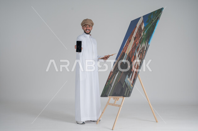 أنشطة ترفيهية فنية ممتعة، استخدام ريشة التلوين، بورتريه لرسام عربي خليجي سعودي يرتدي الثوب التقليدي وقبعة يحمل بيديه هاتف محمول بشاشة سوداء مع الالوان المائية ينظر إلى الكاميرا بإيماءات الفرح و السرور, ورشة عمل وفنون,  صورة الجسم بالطول الكامل, خلفية بيضاء