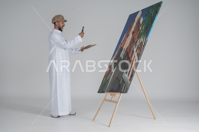 أنشطة ترفيهية ممتعة، استخدام ريشة التلوين، بورتريه لرسام عربي خليجي سعودي يرتدي الثوب التقليدي وقبعة يقوم بالتقاط صورة للوحته الفنية من هاتفه المحمول, ورشة عمل وفنون, صورة الجسم بالطول الكامل,  خلفية بيضاء