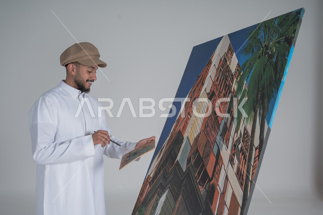 استخدام ريشة التلوين، أنشطة ترفيهية فنية ممتعة، بورتريه مقرب لرسام عربي خليجي سعودي يرتدي الثوب التقليدي وقبعة يحمل بيديه  الالوان المائية, ورشة عمل وفنون,  خلفية بيضاء