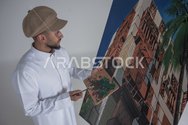 استخدام ريشة التلوين، أنشطة ترفيهية فنية ممتعة، بورتريه مقرب لرسام عربي خليجي سعودي يرتدي الثوب التقليدي وقبعة يحمل بيديه  الالوان المائية, ورشة عمل وفنون,  خلفية بيضاء