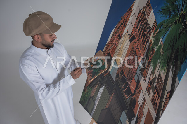 استخدام ريشة التلوين، أنشطة ترفيهية فنية ممتعة، بورتريه مقرب لرسام عربي خليجي سعودي يرتدي الثوب التقليدي وقبعة يحمل بيديه  الالوان المائية, ورشة عمل وفنون,  خلفية بيضاء