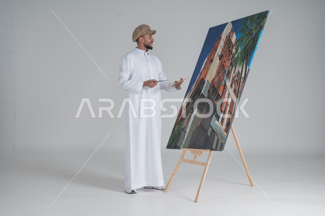ورشة عمل وفنون, استخدام ريشة التلوين، أنشطة ترفيهية فنية ممتعة، بورتريه لرسام عربي خليجي سعودي يرتدي الثوب التقليدي وقبعة يحمل بيديه  الالوان المائية, صورة الجسم بالطول الكامل, خلفية بيضاء