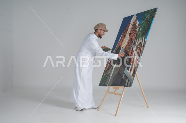 ورشة عمل وفنون, استخدام ريشة التلوين، أنشطة ترفيهية فنية ممتعة، بورتريه لرسام عربي خليجي سعودي يرتدي الثوب التقليدي وقبعة يحمل بيديه  الالوان المائية, صورة الجسم بالطول الكامل, خلفية بيضاء