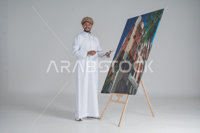 ورشة عمل وفنون, استخدام ريشة التلوين، أنشطة ترفيهية فنية ممتعة، بورتريه لرسام عربي خليجي سعودي يرتدي الثوب التقليدي وقبعة يحمل بيديه  الالوان المائية وينظر الى الكاميرا بايماءات السعادة والسرور, صورة الجسم بالطول الكامل, خلفية بيضاء