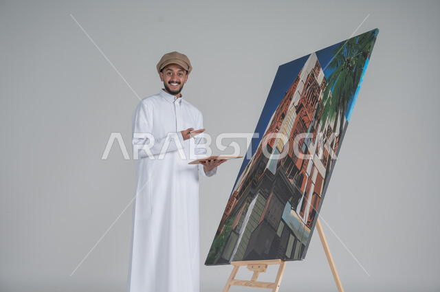ورشة عمل وفنون, استخدام ريشة التلوين، أنشطة ترفيهية فنية ممتعة، بورتريه لرسام عربي خليجي سعودي يرتدي الثوب التقليدي وقبعة يحمل بيديه  الالوان المائية وينظر الى الكاميرا بايماءات السعادة والسرور, خلفية بيضاء