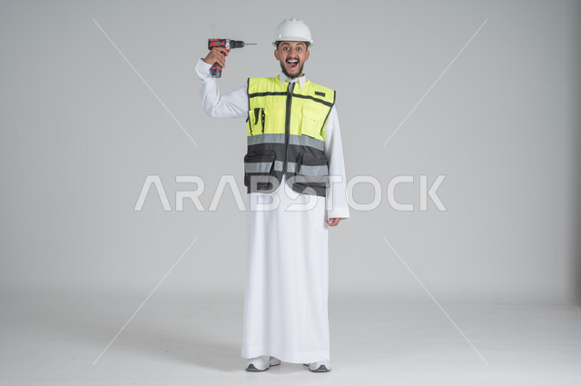 Using maintenance tools and equipment to carry out repairs, skill and mastery of work, portrait of a Saudi Gulf Arab man wearing a thobe and jacket and wearing a protective helmet, holding a pen in his hand and directing it towards his head, full-length body portrait, white background.