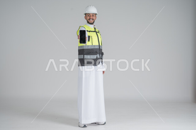 تعابير السعادة والسرور, استخدام التقنيات الحديثة لتسهيل القيام بمهام العمل اليومية، بورتريه لرجل عربي سعودي خليجي يرتدي الثوب والسترة ويضع خوذه الحماية ويحمل بيده هاتف محمول بشاشة سوداء,  صورة الجسم بالطول الكامل, خلفية بيضاء