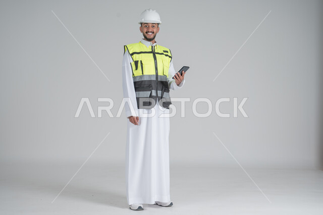 Using modern technologies, following up on work and tasks, supervising and controlling engineering projects, a portrait of a Saudi Gulf Arab man wearing a thobe and jacket, wearing a protective helmet and holding a mobile phone in his hand, full-length body portrait, white background