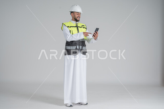 استخدام التقنيات الحديثة، متابعة العمل والمهام، الاشراف والرقابة على المشاريع الهندسية، بورتريه لرجل عربي سعودي خليجي يرتدي الثوب والسترة ويضع خوذه الحماية ويحمل بيده الهاتف المحمول,  صورة الجسم بالطول الكامل, خلفية بيضاء