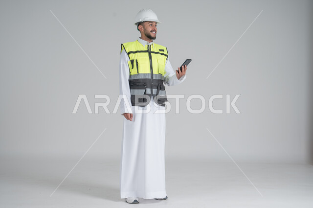 Using modern technologies, following up on work and tasks, supervising and controlling engineering projects, a portrait of a Saudi Gulf Arab man wearing a thobe and jacket, wearing a protective helmet and holding a mobile phone in his hand, full-length body portrait, white background