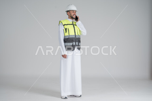 استخدام التقنيات الحديثة، متابعة العمل والمهام، الاشراف والرقابة على المشاريع الهندسية، بورتريه لرجل عربي سعودي خليجي يرتدي الثوب والسترة ويضع خوذه الحماية ويحمل بيده الهاتف المحمول,  صورة الجسم بالطول الكامل, خلفية بيضاء