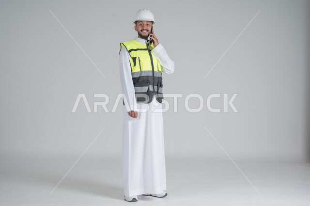 استخدام التقنيات الحديثة، متابعة العمل والمهام، الاشراف والرقابة على المشاريع الهندسية، بورتريه لرجل عربي سعودي خليجي يرتدي الثوب والسترة ويضع خوذه الحماية ويحمل بيده الهاتف المحمول,  صورة الجسم بالطول الكامل, خلفية بيضاء