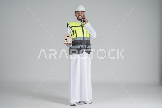 Gestures of happiness and pleasure, using modern technologies, following up on work and tasks, supervising and controlling engineering projects, a portrait of a Saudi Gulf Arab man wearing a dress and jacket, wearing a protective helmet, holding a mobile phone and a model of a residential house in his hands, full-length body portrait, white background.