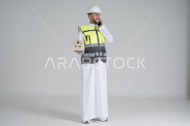 ايماءات السعادة والسرور, استخدام التقنيات الحديثة، متابعة العمل والمهام، الاشراف والرقابة على المشاريع الهندسية، بورتريه لرجل عربي سعودي خليجي يرتدي الثوب والسترة ويضع خوذه الحماية ويحمل بيديه الهاتف المحمول ومجسم منزل سكني,  صورة الجسم بالطول الكامل, خلفية بيضاء