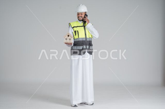 ايماءات السعادة والسرور, استخدام التقنيات الحديثة، متابعة العمل والمهام، الاشراف والرقابة على المشاريع الهندسية، بورتريه لرجل عربي سعودي خليجي يرتدي الثوب والسترة ويضع خوذه الحماية ويحمل بيديه الهاتف المحمول ومجسم منزل سكني,  صورة الجسم بالطول الكامل, خلفية بيضاء