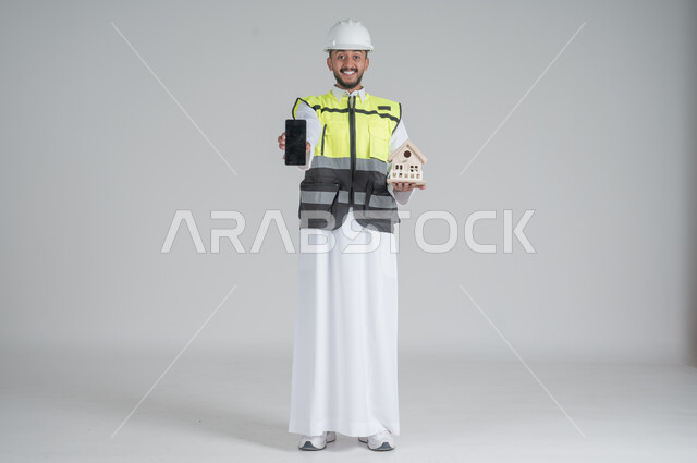 ايماءات السعادة والسرور, استخدام التقنيات الحديثة، متابعة العمل والمهام، الاشراف والرقابة على المشاريع الهندسية، بورتريه لرجل عربي سعودي خليجي يرتدي الثوب والسترة ويضع خوذه الحماية ويحمل بيديه هاتف محمول بشاشة سوادء ومجسم لمنزل سكني,  صورة الجسم بالطول الكامل, خلفية بيضاء