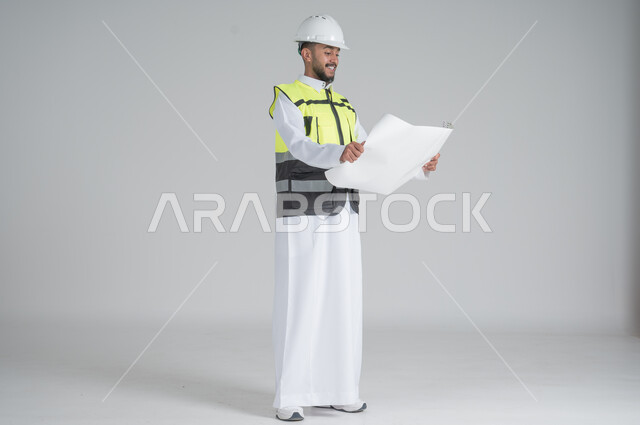 ايماءات السعادة والسرور, متابعة الأعمال الهندسية, النظر إلى مخطط الإنشاءات, مهن ووظائف سعودية, تنمية وتطوير المجال الهندسي, بورتريه لرجل عربي سعودي خليجي يرتدي الثوب والسترة ويضع خوذه الحماية , صورة الجسم بالطول الكامل, خلفية بيضاء