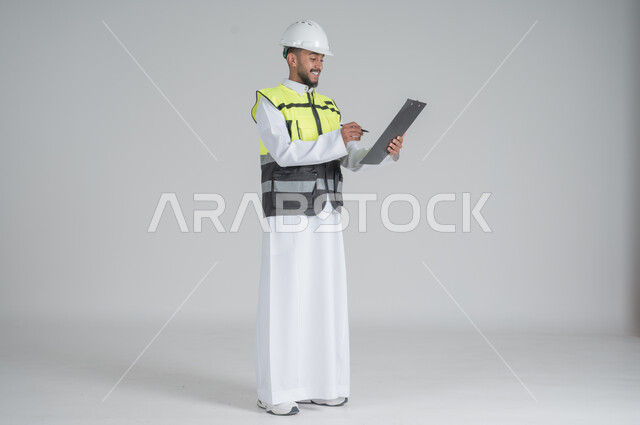 ايماءات السعادة والسرور, متابعة الأعمال الهندسية, مهن ووظائف سعودية, تنمية وتطوير المجال الهندسي, بورتريه لرجل عربي سعودي خليجي يرتدي الثوب والسترة ويضع خوذه الحماية, القيام بتسجيل الملاحظات على المخطط، صورة الجسم بالطول الكامل, خلفية بيضاء