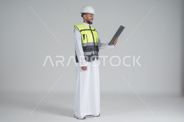 ايماءات السعادة والسرور, متابعة الأعمال الهندسية, مهن ووظائف سعودية, تنمية وتطوير المجال الهندسي, بورتريه لرجل عربي سعودي خليجي يرتدي الثوب والسترة ويضع خوذه الحماية, القيام بتسجيل الملاحظات على المخطط، صورة الجسم بالطول الكامل, خلفية بيضاء