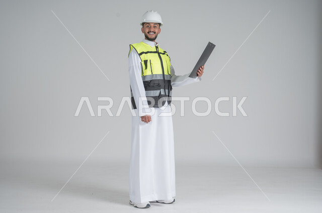 ايماءات السعادة والسرور, متابعة الأعمال الهندسية, مهن ووظائف سعودية, تنمية وتطوير المجال الهندسي, بورتريه لرجل عربي سعودي خليجي يرتدي الثوب والسترة ويضع خوذه الحماية, القيام بتسجيل الملاحظات على المخطط، صورة الجسم بالطول الكامل, خلفية بيضاء