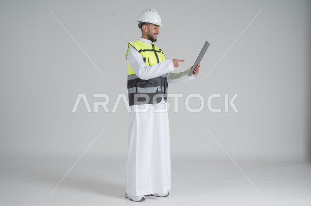 ايماءات السعادة والسرور, متابعة الأعمال الهندسية, مهن ووظائف سعودية, تنمية وتطوير المجال الهندسي, بورتريه لرجل عربي سعودي خليجي يرتدي الثوب والسترة ويضع خوذه الحماية, القيام بتسجيل الملاحظات على المخطط، صورة الجسم بالطول الكامل, خلفية بيضاء