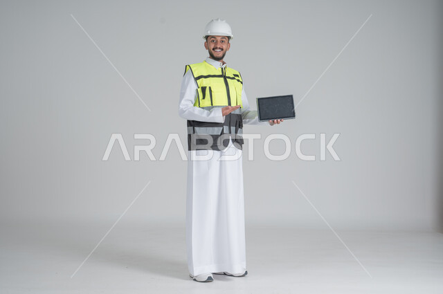 الإشارة إلى شاشة سوداء فارغة, استخدام التابلت والتقنيات الحديثة لتسهيل القيام بمهام العمل اليومية، مهن ووظائف سعودية, تنمية وتطوير المجال الهندسي, بورتريه لرجل عربي سعودي خليجي يرتدي الثوب والسترة ويضع خوذه الحماية, صورة الجسم بالطول الكامل, خلفية بيضاء