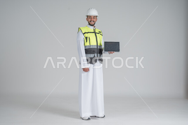 استخدام التقنيات الحديثة لتسهيل القيام بمهام العمل اليومية، مهن ووظائف سعودية, تنمية وتطوير المجال الهندسي, بورتريه لرجل عربي سعودي خليجي يرتدي الثوب والسترة ويضع خوذه الحماية يحمل بيده جهاز تابليت بشاشة سوداء, صورة الجسم بالطول الكامل, خلفية بيضاء