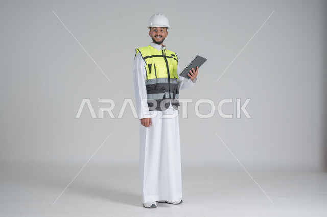 ايماءات السعادة والسرور استخدام التقنيات الحديثة لتسهيل القيام بمهام العمل اليومية، مهن ووظائف سعودية, تنمية وتطوير المجال الهندسي, بورتريه لرجل عربي سعودي خليجي يرتدي الثوب والسترة ويضع خوذه الحماية يحمل بيده جهاز تابليت, صورة الجسم بالطول الكامل, خلفية بيضاء