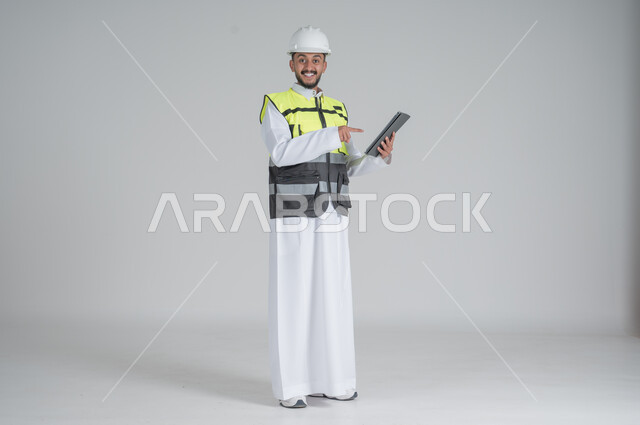 ايماءات السعادة والسرور استخدام التقنيات الحديثة لتسهيل القيام بمهام العمل اليومية، مهن ووظائف سعودية, تنمية وتطوير المجال الهندسي, بورتريه لرجل عربي سعودي خليجي يرتدي الثوب والسترة ويضع خوذه الحماية يحمل بيده جهاز تابليت, صورة الجسم بالطول الكامل, خلفية بيضاء