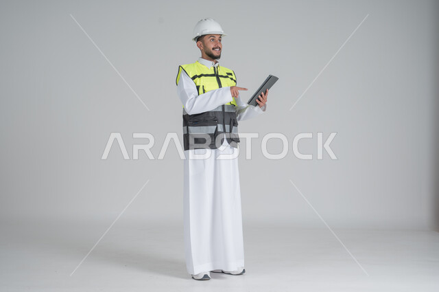 ايماءات السعادة والسرور استخدام التقنيات الحديثة لتسهيل القيام بمهام العمل اليومية، مهن ووظائف سعودية, تنمية وتطوير المجال الهندسي, بورتريه لرجل عربي سعودي خليجي يرتدي الثوب والسترة ويضع خوذه الحماية يحمل بيده جهاز تابليت, صورة الجسم بالطول الكامل, خلفية بيضاء