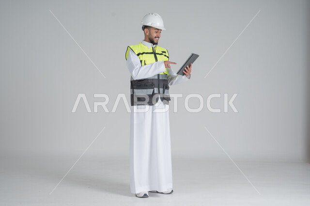 ايماءات السعادة والسرور استخدام التقنيات الحديثة لتسهيل القيام بمهام العمل اليومية، مهن ووظائف سعودية, تنمية وتطوير المجال الهندسي, بورتريه لرجل عربي سعودي خليجي يرتدي الثوب والسترة ويضع خوذه الحماية يحمل بيده جهاز تابليت, صورة الجسم بالطول الكامل, خلفية بيضاء
