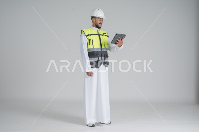 ايماءات السعادة والسرور استخدام التقنيات الحديثة لتسهيل القيام بمهام العمل اليومية، مهن ووظائف سعودية, تنمية وتطوير المجال الهندسي, بورتريه لرجل عربي سعودي خليجي يرتدي الثوب والسترة ويضع خوذه الحماية يحمل بيده جهاز تابليت, صورة الجسم بالطول الكامل, خلفية بيضاء